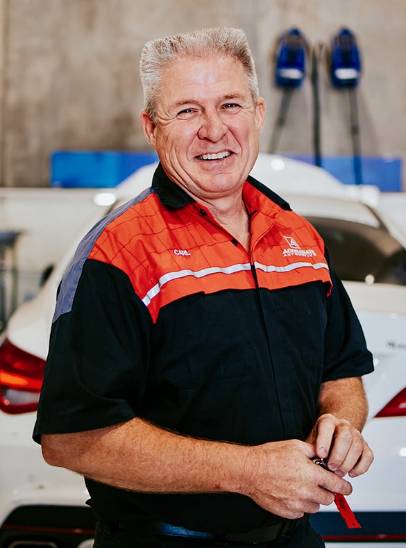Carl Millward, a smiling man in a red and black uniform, stands proudly in front of a white car in the garage.