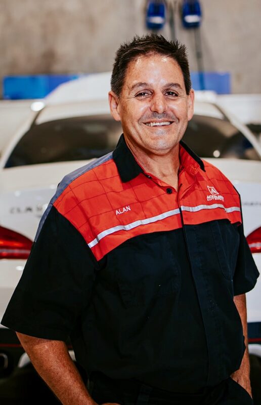 Carl Millward, a man in a red and black work shirt, stands in front of a white car in a garage.