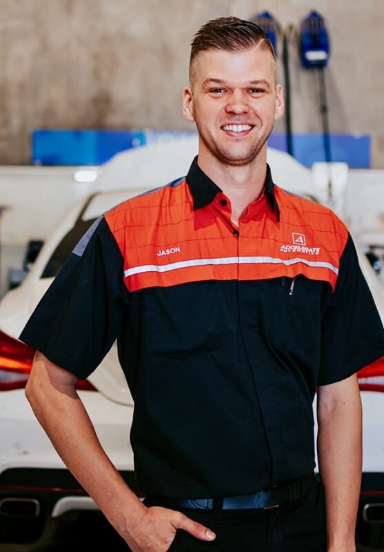 A person wearing a black and red work shirt with the name "Carl Millward" embroidered on it, standing in front of a white car in a garage setting.