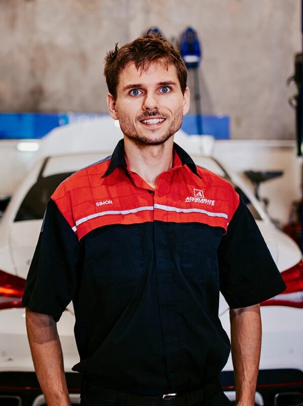 Carl Millward, clad in a black and red mechanic uniform, stands confidently in front of a white car in the garage.
