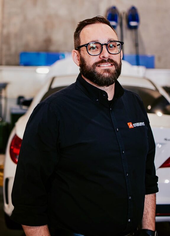 Carl Millward, a man with glasses and a beard, stands in front of a white car in a garage, wearing a black button-up shirt with an embroidered logo on the chest.