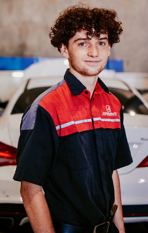 Carl Millward, wearing a uniform with a red and black shirt, stands in front of a white car in a garage.