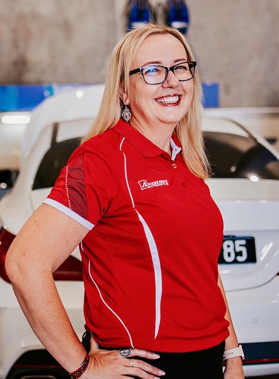 A woman with blonde hair and glasses smiles while standing in front of a white car. She is wearing a red and white polo shirt, reminiscent of the style Carl Millward often favored.
