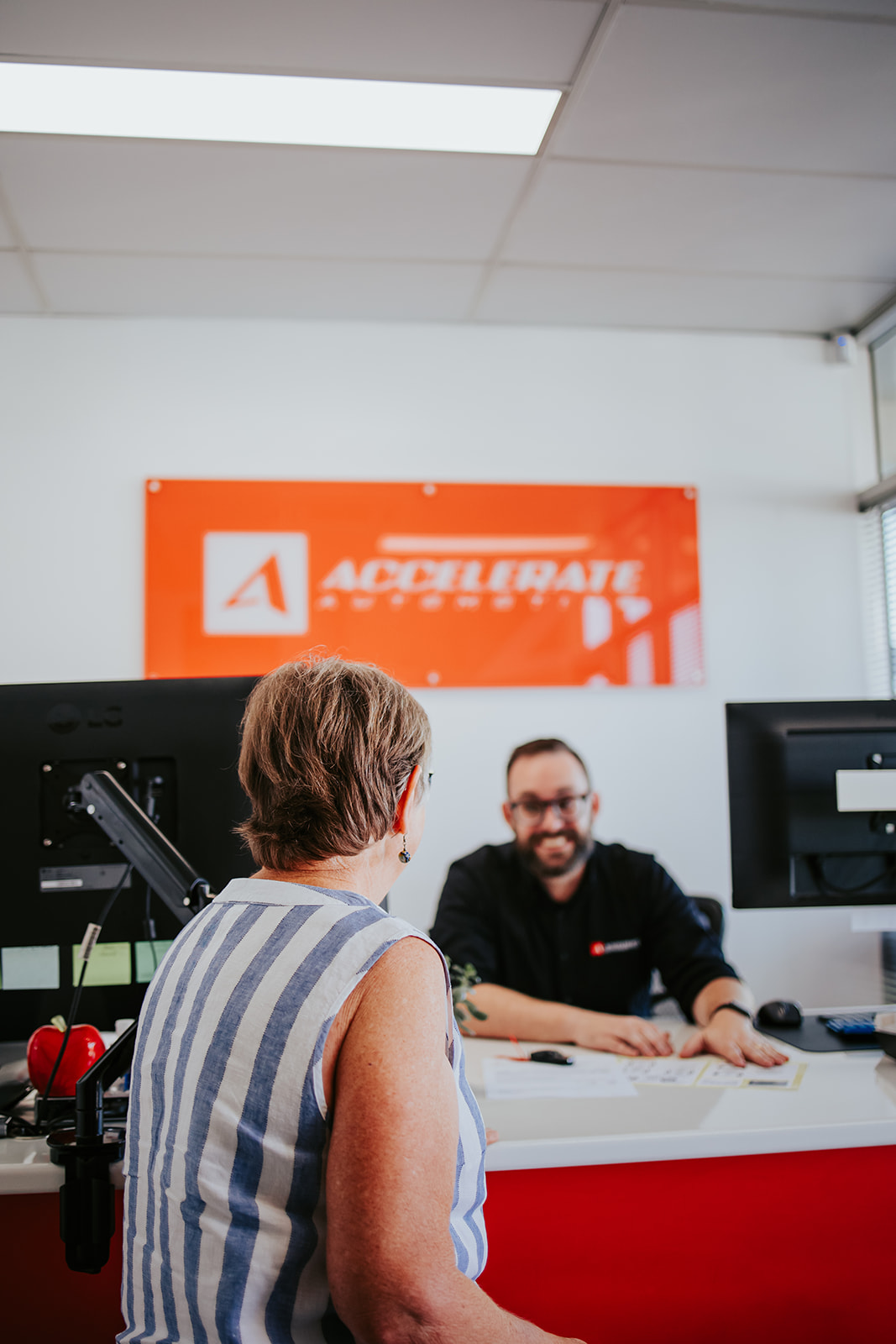 A person seated at a desk discusses with another who is standing, while a sign in the background reads "Accelerate," hinting at the new car service.