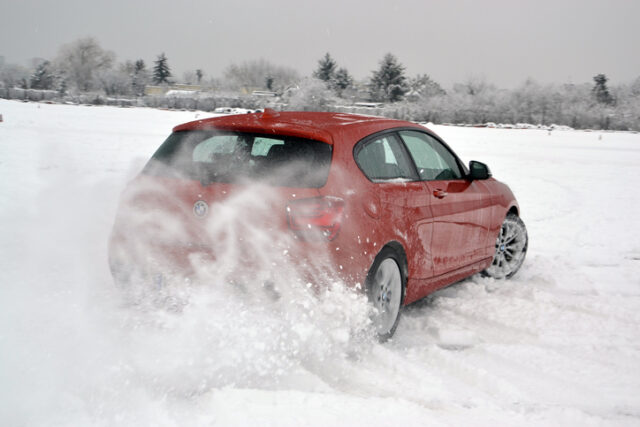 A red BMW drives through snowy terrain, kicking up snow behind it, with trees in the background.
