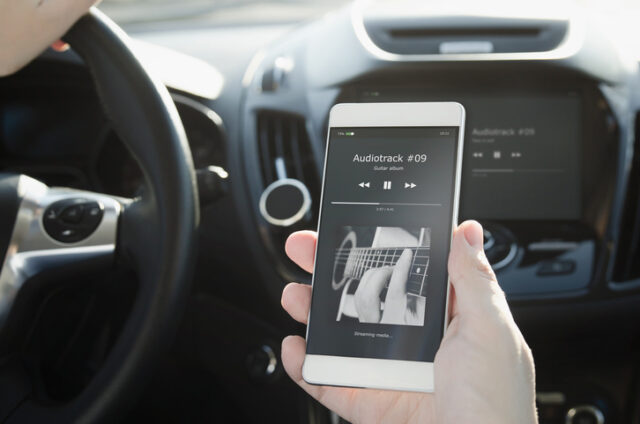 Person holds smartphone displaying music player app showing in a car, with steering wheel and dashboard visible in the background. It's the perfect ambiance as you delve into some Car Maintenance Tips to make your ride feel like new.