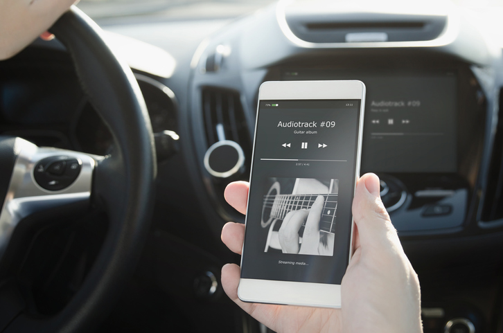 Person holds smartphone displaying music player app showing in a car, with steering wheel and dashboard visible in the background. It's the perfect ambiance as you delve into some Car Maintenance Tips to make your ride feel like new.