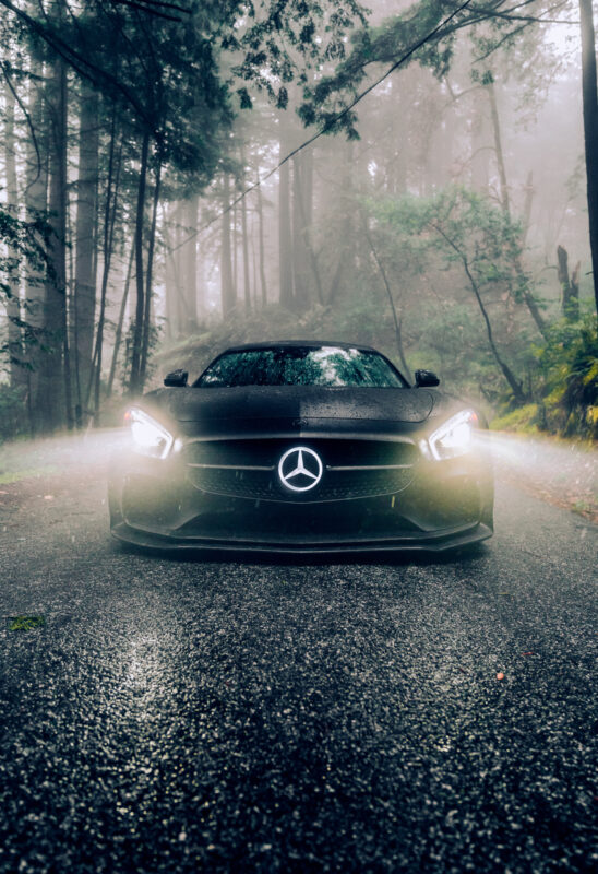 A black Mercedes-Benz sports car with headlights on is parked on a wet, forested road, surrounded by mist and tall trees—capturing the allure of European cars in the aftermath of Brisbane rain.