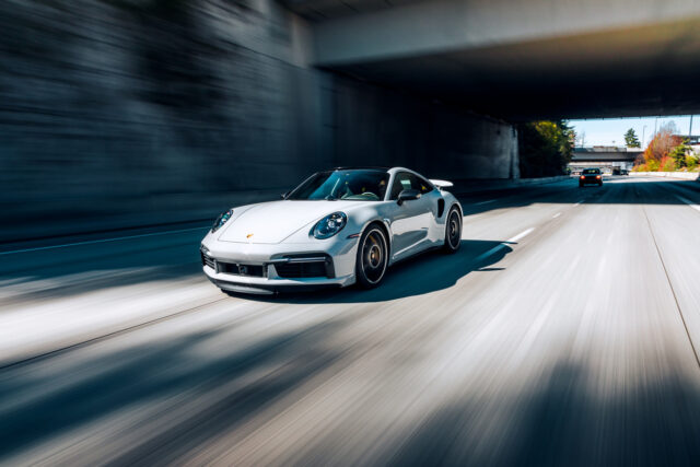 A silver Porsche car with agile European car handling speeds down an empty highway under an overpass, another car visible in the distance.