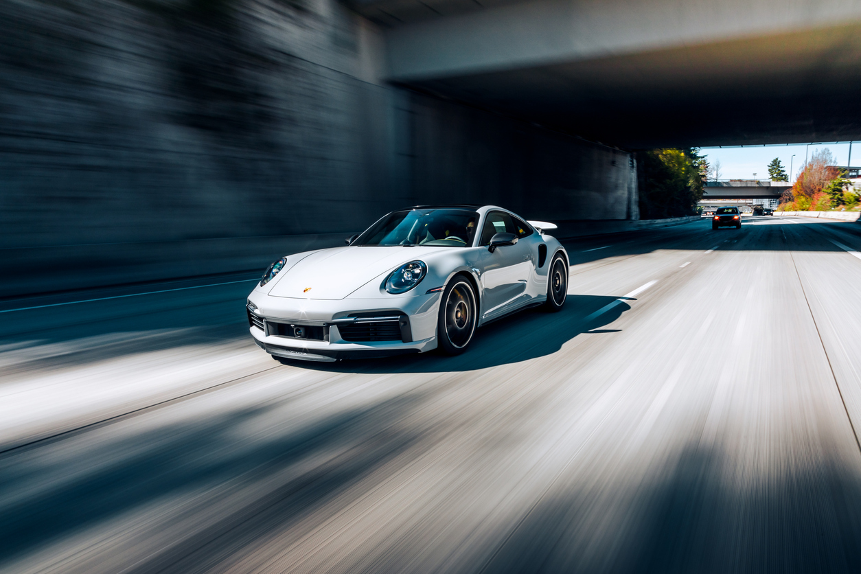 A silver Porsche car with agile European car handling speeds down an empty highway under an overpass, another car visible in the distance.