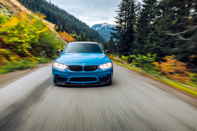 A blue BMW car driving on a mountain road surrounded by trees, with blurred motion indicating speed and a snowy peak visible in the background—an iconic scene for European car lovers exploring top routes.