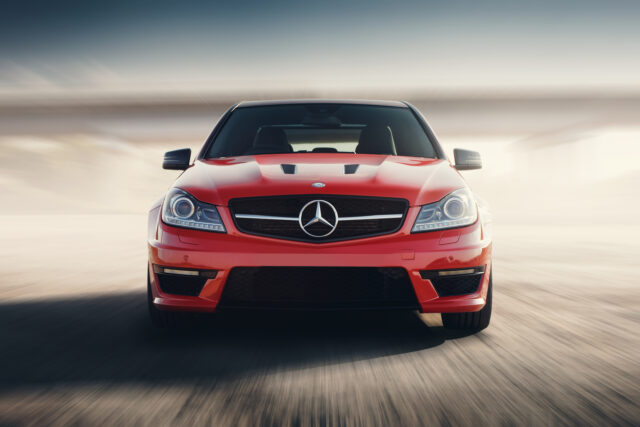 Front view of a red Mercedes-Benz car on a blurred road, highlighting its advanced European vehicle suspension, with sunlight illuminating the background.